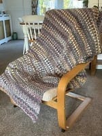 A large, textured crochet blanket featuring variegated neutral shades of brown, tan, and cream, draped over a light wooden armchair.