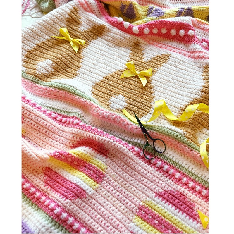 Close-up of a crocheted Easter blanket featuring brown bunny motifs with yellow bows and colorful striped eggs.